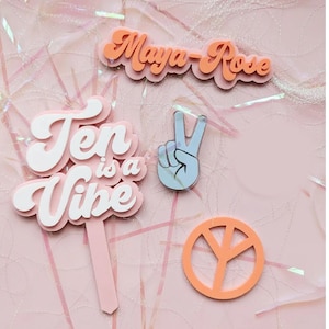 May include: A set of four cake toppers in pink, white and orange. The toppers feature the text "Ten is a Vibe", a peace sign, a hand making the peace sign, and the name "Maya-Rose".