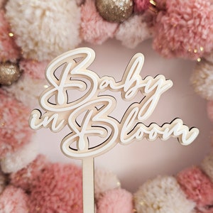 Pode incluir: Um letreiro de madeira branco com o texto "Baby in Bloom" em uma fonte cursiva. O letreiro está preso a um bastão de madeira e é exibido na frente de uma coroa de pompons rosa e brancos.