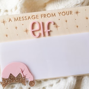 Puede incluir: Cartel de madera con el texto "A MESSAGE FROM YOUR ELF" y un espacio en blanco para un mensaje. La palabra "elf" es rosa, y un gorro de elfo rosa con purpurina dorada está en la parte inferior.