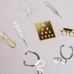 May include: A set of silver and gold medical themed decorations. The set includes two stethoscopes, two syringes, a pair of glasses, and an eye chart. The eye chart is gold and has the letters E, FP, TOZ, and LPED on it.