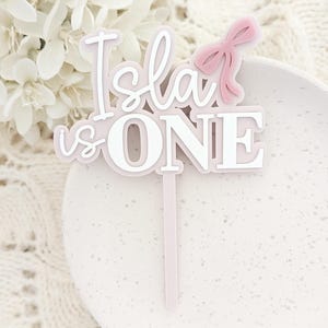 May include: Pink and white acrylic cake topper with the text "Isla is ONE" and a pink bow.