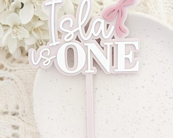 May include: Pink and white acrylic cake topper with the text "Isla is ONE" and a pink bow.