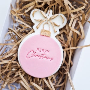 Peut inclure: Un biscuit en forme d'ornement de Noël rose et blanc avec un nœud doré et l'inscription "Merry Christmas" en lettres rouges.