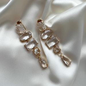 Pode incluir: Um par de brincos de argola de tom dourado com pedras de cristal facetadas transparentes. Os brincos são longos e apresentam uma cascata de pedras.