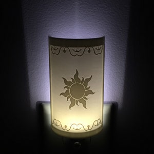 May include: A white night light with a sun design on the front. The sun is stylized with a yellow center and white rays. The night light is plugged into an outlet.