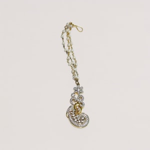 May include: A gold-toned headpiece with a dangling pendant. The pendant features a floral design and a curved, embellished shape, encrusted with clear crystals. A strand of small, white pearls connects the pendant to a hook.