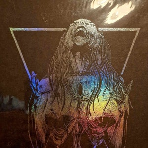May include: A black and white illustration of a figure with long hair, open mouth, and raised arms, inside a triangle. The figure is surrounded by a holographic iridescent effect. A skull is at the bottom of the triangle.