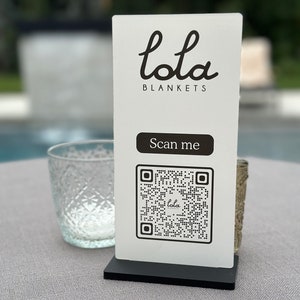 May include: White sign with black text and a QR code. The text reads "lola BLANKETS" and "Scan me". The QR code has the word "lola" in the center.