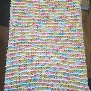 May include: A chunky knit blanket in a pastel color palette of yellow, pink, blue, and white. The blanket is made with a thick yarn and has a soft, fluffy texture.