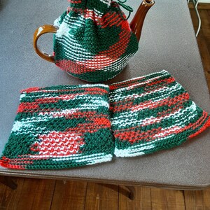 Op de afbeelding: Een set van drie gebreide theepot-hoezen in groen, wit en rood ruitjespatroon. De hoezen zijn gemaakt van een dik garen en hebben een eenvoudig, klassiek ontwerp.