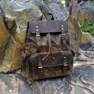 May include: A brown leather backpack with multiple straps and buckles. The backpack has a distressed look and is sitting on a rocky surface.