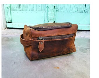 May include: A brown leather travel bag with a zipper closure and a handle. The bag is made of distressed leather and has a rustic look.