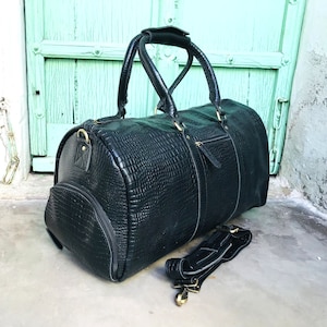 May include: A black leather duffel bag with a textured crocodile pattern. The bag has two top handles and a detachable shoulder strap.