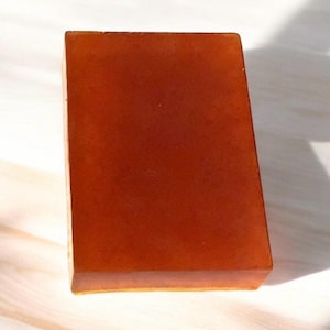 May include: A rectangular bar of translucent amber-coloured soap with smooth, flat surfaces and sharp, defined edges. The soap has a uniform, deep orange-brown hue throughout its solid, semi-transparent body, reflecting light on its top surface.