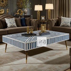 May include: A white and blue striped coffee table with a geometric pattern inlay. The table has four gold metal legs and is sitting on a cream-colored rug.