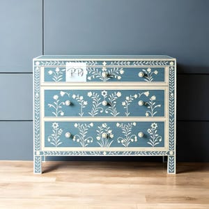 以下が含まれることがあります： 青い塗装仕上げと白い花の象嵌が施された3段引き出しのチェスト。チェストには白いノブの引き出しが3つあり、上部と下部に白い縁取りが施されています。
