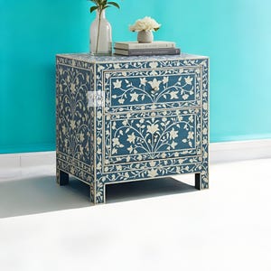 Puede incluir: Una mesita de noche azul con dos cajones, con intrincados dise&ntilde;os florales blancos de incrustaciones de hueso. La mesita de noche tiene un estilo moderno y es perfecta para un dormitorio o una sala de estar.
