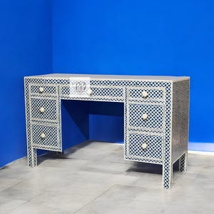 Op de afbeelding: Een witte bureau met een blauw geometrisch patroon en meerdere lades. De bureau heeft een rechthoekige vorm met een platte bovenkant en wordt ondersteund door vier poten. De lades hebben ronde witte knoppen.