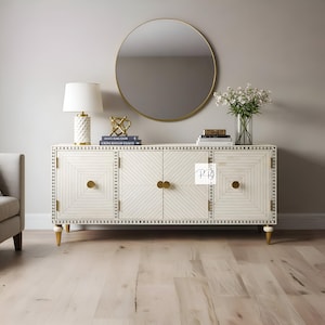 Può includere: Un credenza bianca con intarsi in osso con tre ante e ferramenta dorata. La credenza presenta un motivo geometrico e si trova su un pavimento in legno chiaro. C'è uno specchio rotondo, una lampada e un vaso di fiori sopra la credenza.