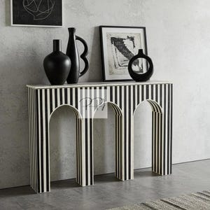 Könnte beinhalten: Eine Konsole mit schwarz-weißen Streifen und einem bogenförmigen Sockel. Der Tisch ist mit schwarzen Vasen und gerahmten Kunstwerken versehen. Das Design weist vertikale Streifen auf, die ein kühnes, grafisches Statement für die Inneneinrichtung bilden.