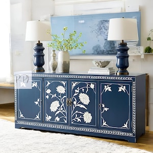 May include: A blue wooden cabinet with four doors and white floral inlays. The cabinet has a white top and is decorated with a white trim. Two white lamps with white shades are on top of the cabinet.