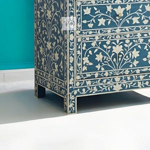 Puede incluir: Un cofre de cajones azul con incrustaciones florales blancas. El cofre tiene dos cajones y se apoya en cuatro patas.