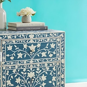 Puede incluir: Una mesita de noche de madera azul con dos cajones, con intrincados dise&ntilde;os de incrustaciones florales blancas. La mesita de noche tiene una flor blanca en la parte superior y dos libros con los t&iacute;tulos "Serpent in the Water" y "Continuum Cairo Raw Tonic Canary".