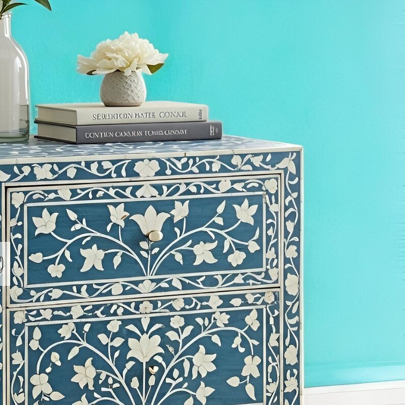 Puede incluir: Una mesita de noche de madera azul con dos cajones, con intrincados dise&ntilde;os de incrustaciones florales blancas. La mesita de noche tiene una flor blanca en la parte superior y dos libros con los t&iacute;tulos "Serpent in the Water" y "Continuum Cairo Raw Tonic Canary".