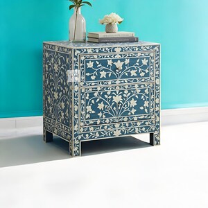 Puede incluir: Una mesita de noche azul con dos cajones, con intrincados dise&ntilde;os florales blancos de incrustaciones de hueso. La mesita de noche tiene un estilo moderno y es perfecta para un dormitorio o una sala de estar.