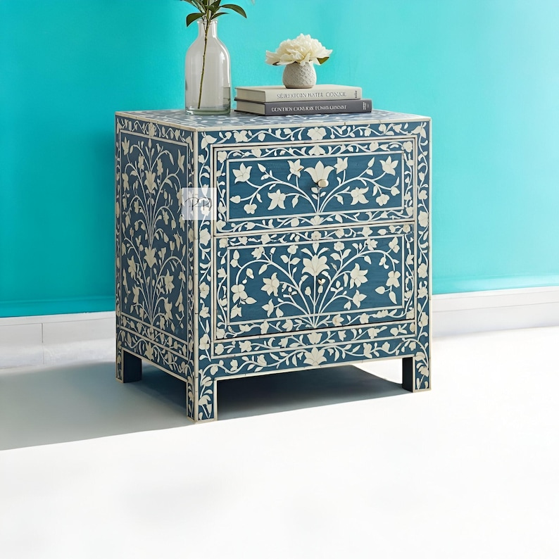 Puede incluir: Una mesita de noche azul con dos cajones, con intrincados dise&ntilde;os florales blancos de incrustaciones de hueso. La mesita de noche tiene un estilo moderno y es perfecta para un dormitorio o una sala de estar.