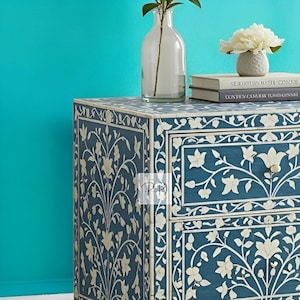 Puede incluir: Una mesita de noche de madera azul con dos cajones presenta intrincados dise&ntilde;os florales blancos en incrustaciones. La mesita de noche est&aacute; decorada con un jarr&oacute;n de flores y libros.