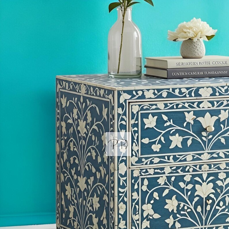Puede incluir: Una mesita de noche de madera azul con dos cajones presenta intrincados dise&ntilde;os florales blancos en incrustaciones. La mesita de noche est&aacute; decorada con un jarr&oacute;n de flores y libros.