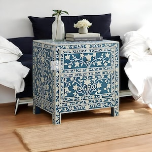 Puede incluir: Una mesita de noche azul de dos cajones con incrustaciones florales blancas. La mesita de noche est&aacute; hecha de madera y tiene un dise&ntilde;o tradicional. Est&aacute; sentada sobre una alfombra marr&oacute;n claro frente a un sof&aacute;.