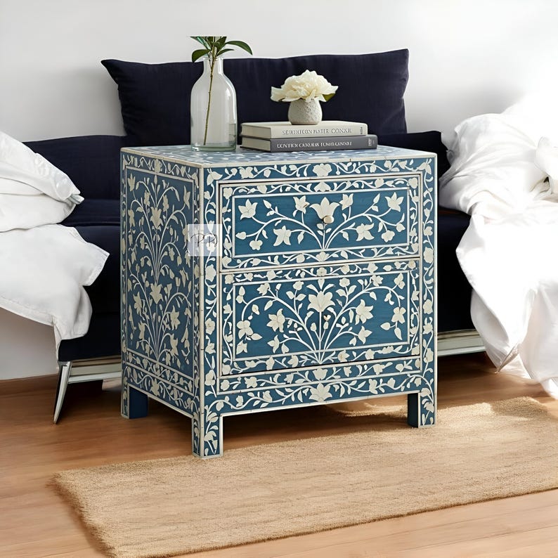 Puede incluir: Una mesita de noche azul de dos cajones con incrustaciones florales blancas. La mesita de noche est&aacute; hecha de madera y tiene un dise&ntilde;o tradicional. Est&aacute; sentada sobre una alfombra marr&oacute;n claro frente a un sof&aacute;.