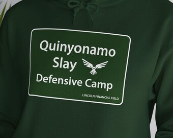 May include: A dark green hoodie featuring a white graphic of a sign that reads "Quinnyonamo Slay Defensive Camp" with a bird logo and the text "Lincoln Financial Field" underneath.