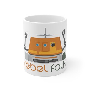 May include: White ceramic mug featuring a cartoon illustration of a droid with orange, grey, and blue details. The text "rebel folk" is printed in green and orange at the bottom of the mug.
