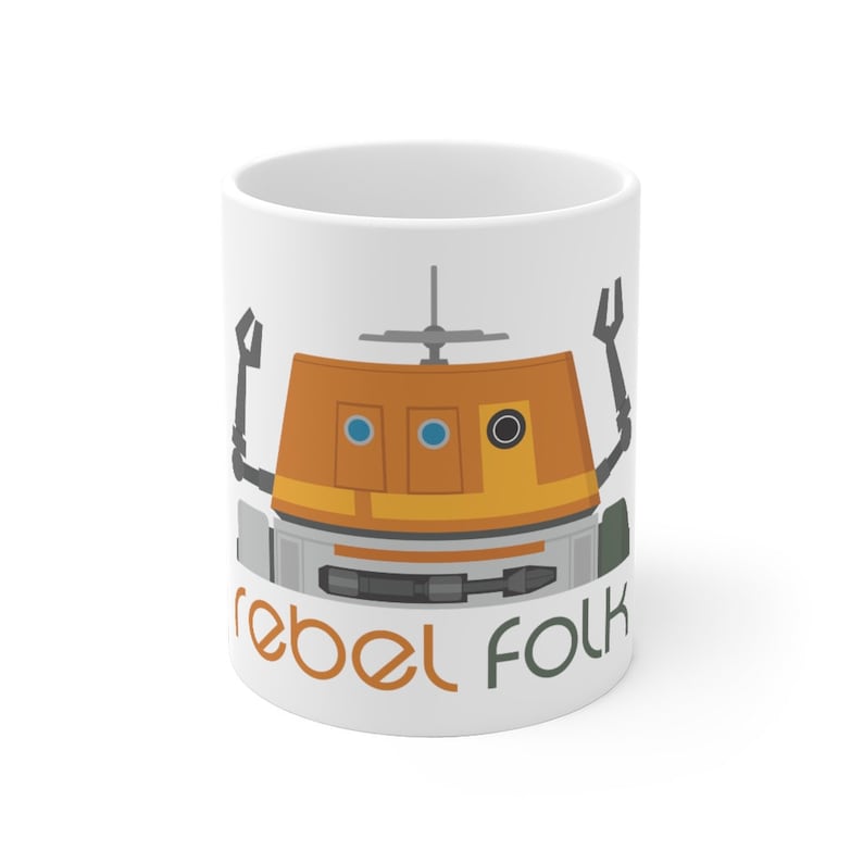 May include: White ceramic mug featuring a cartoon illustration of a droid with orange, grey, and blue details. The text "rebel folk" is printed in green and orange at the bottom of the mug.