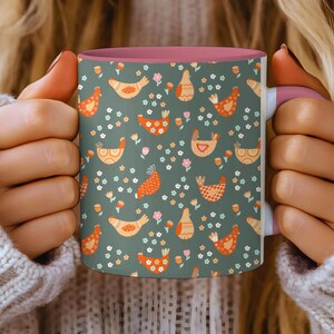 May include: A ceramic mug with a pink interior and handle, featuring a repeating pattern of orange and yellow chickens, small flowers, and leaves on a dark green background. The mug is held by a person wearing a white sweater.