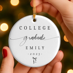 Puede incluir: Un adorno navideño de cerámica blanca con una cuerda dorada, sostenido por manos con esmalte de uñas rojo. El adorno tiene las palabras "COLLEGE graduate EMILY -2025-" en escritura negra y un lazo.