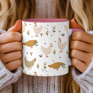 Puede incluir: Taza de cerámica blanca con interior y asa rosa, con un estampado repetido de pollos de dibujos animados, flores y hojas. La taza la sostiene una persona que lleva un suéter blanco.