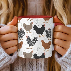 May include: A white ceramic mug with a red interior, featuring a repeating pattern of various chicken illustrations in brown, black, and speckled designs. The mug is held by a person wearing a white sweater.