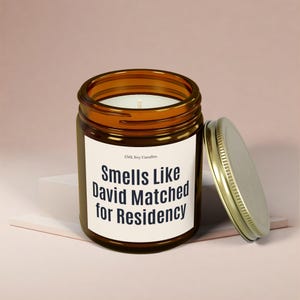 Puede incluir: Un tarro de vela de vidrio marrón con una etiqueta blanca que dice "Smells Like David Matched for Residency". El tarro de vela tiene una tapa dorada.