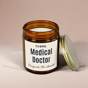 Puede incluir: Un tarro de vela de vidrio marrón con una etiqueta blanca que dice "It is giving, Medical Doctor Congrats Dr. Smith!" La tapa es dorada y está abierta.