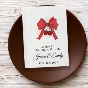 Op de afbeelding: Een witte kaart met een rode strik en twee kersen erop. De kaart zegt "Advies voor de toekomstige ouders Jason & Emily EST. JUNI 2025".