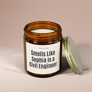 Puede incluir: Un tarro de vela de vidrio marrón con una etiqueta blanca que dice "Smells Like Sophia is a Civil Engineer". El tarro de vela tiene una tapa dorada.