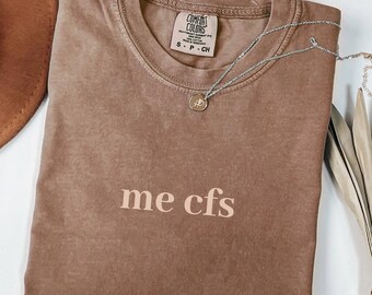 May include: A brown t-shirt with the text "me cfs" printed in a light pink color. The shirt is made of a soft, comfortable material.