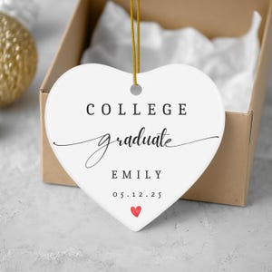 Puede incluir: Un adorno blanco en forma de corazón con las palabras "COLLEGE graduate EMILY 05.12.25" en texto negro. Un pequeño corazón rojo está en la parte inferior. El adorno cuelga de una cuerda dorada y está dentro de una caja de cartón.