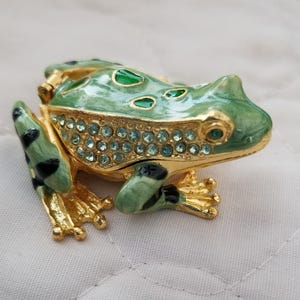 May include: A decorative frog figurine with a hinged lid. The frog is painted in shades of green and gold, with black spots and blue crystal accents. The figurine is made of metal.