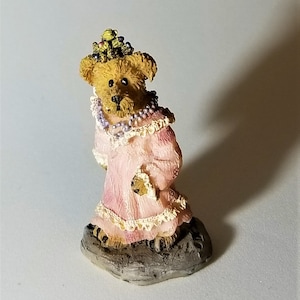 May include: A small, brown teddy bear figurine wearing a pink dress with lace trim and a crown of green leaves. The bear is standing on a gray rock base.