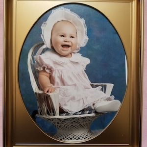 May include: A framed photograph of a baby girl wearing a white bonnet and a pink dress, sitting in a white wicker chair. The baby is smiling and looking at the camera.